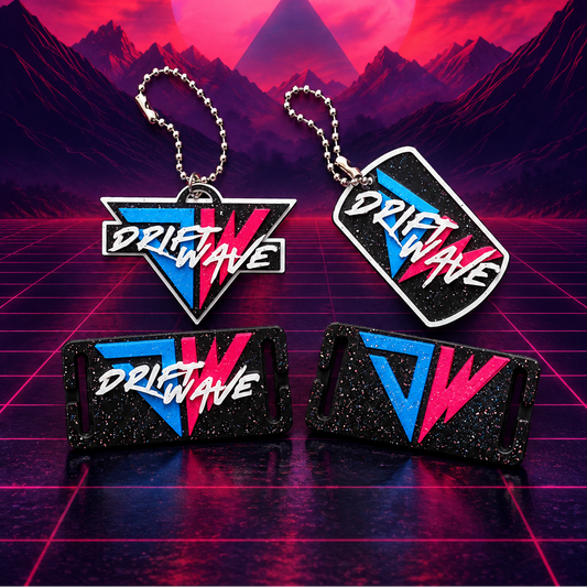 Four Driftwave branded accessories arranged on a neon grid surface with a retro synthwave mountain landscape and large pink sun in the background. The set includes two keychains and two rectangular items, all featuring black glitter or textured surfaces with blue and pink graphic elements and white Driftwave text.
